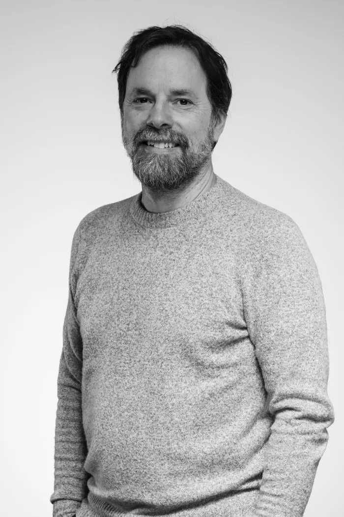 Un homme aux cheveux bruns mi-longs et à la barbe sourit à l'appareil photo. Il porte un pull à manches longues de couleur claire et se tient debout sur un fond clair et uni. La photo est en noir et blanc.