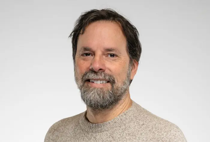 Un homme d'âge moyen aux cheveux bruns et à la barbe sourit à l'appareil photo. Il porte un pull beige et pose devant un fond gris clair uni.