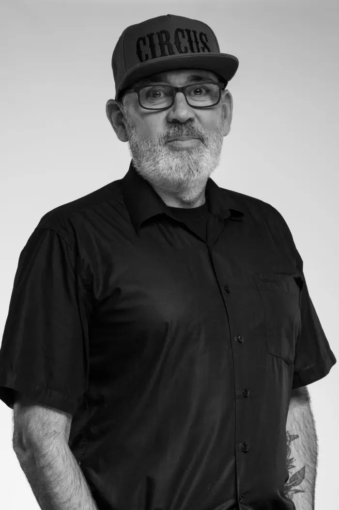 Un homme barbu, portant des lunettes, une casquette de base-ball avec l'inscription CIRCUS et une chemise noire à manches courtes se tient face à la caméra sur un fond uni.