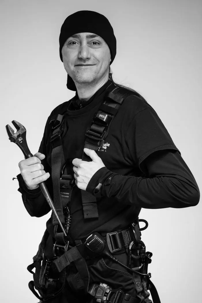 Une personne portant un bonnet noir et un harnais de travail sourit à la caméra, tenant une grosse clé dans une main. Le harnais est équipé de divers outils et équipements. La photo est en noir et blanc.