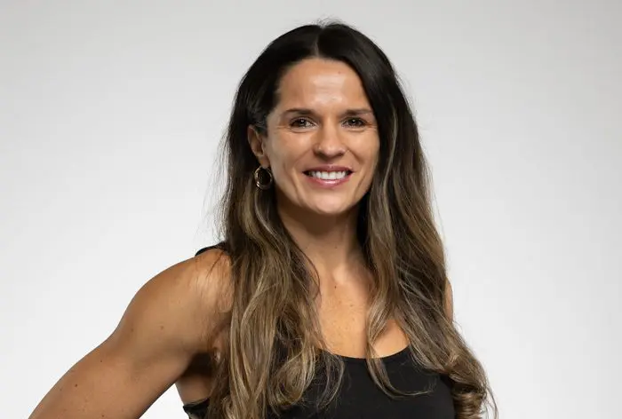 Une femme aux longs cheveux bruns, vêtue d'un haut noir sans manches, sourit à l'appareil photo sur un fond blanc uni.