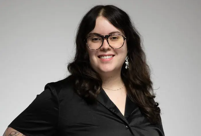 Une personne aux longs cheveux noirs ondulés, portant des lunettes, une chemise noire boutonnée et des boucles d'oreilles pendantes, sourit à l'appareil photo sur un fond gris clair uni.