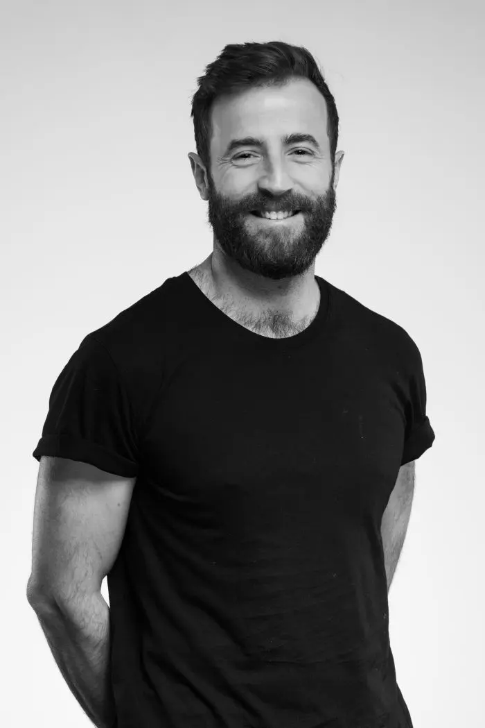 Un homme barbu aux cheveux courts, vêtu d'un t-shirt noir, souriant, debout sur un fond clair et uni, les mains derrière le dos.