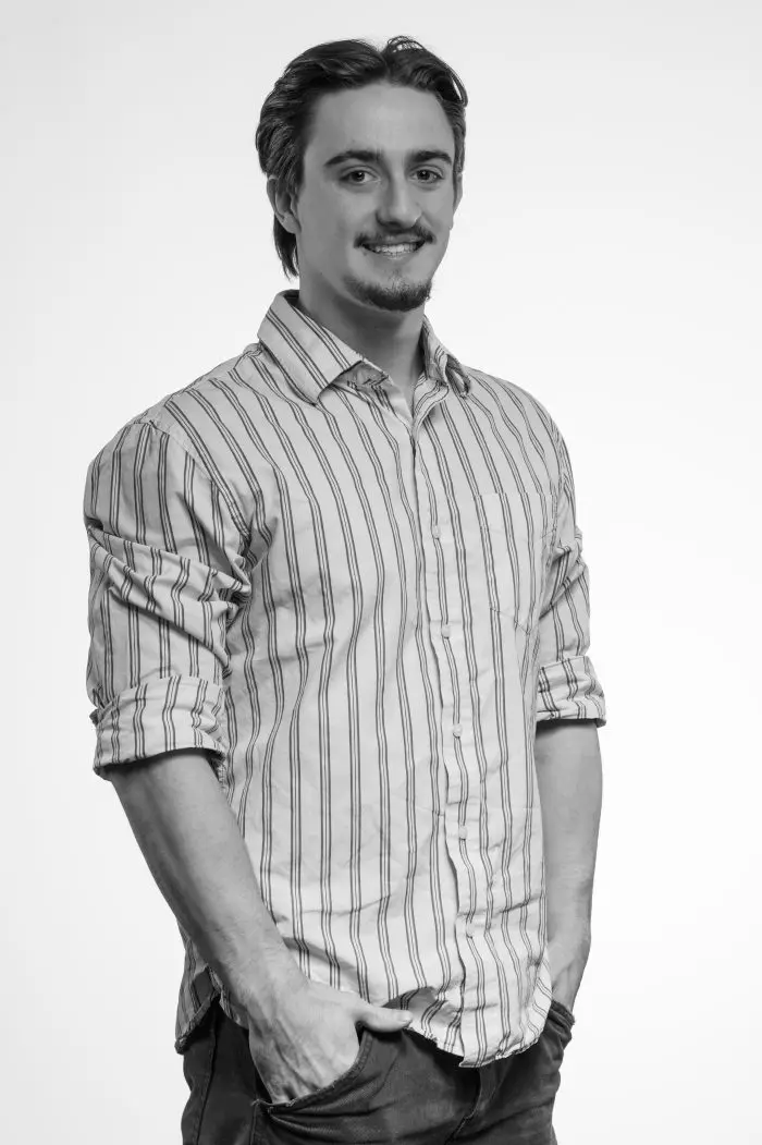 Un jeune homme aux cheveux bruns mi-longs et à la barbichette, vêtu d'une chemise à boutons rayée et d'un jean, se tient debout, les mains dans les poches, souriant à l'appareil photo sur un fond blanc uni.