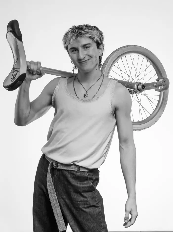 Une personne vêtue d'un débardeur et d'un jean se tient debout, sûre d'elle, tenant un monocycle par-dessus son épaule. La photo est en noir et blanc. La personne a des cheveux courts et clairs et porte des colliers et des boucles d'oreilles.