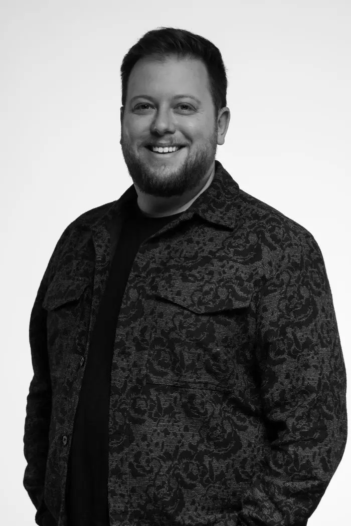 Un homme barbu sourit à la caméra. Il porte une veste sombre à motifs sur une chemise unie. L'arrière-plan est blanc et la photo est en noir et blanc.