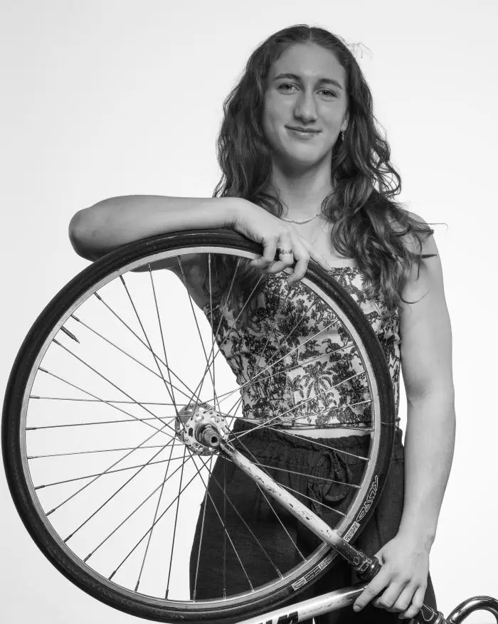 Une jeune femme aux longs cheveux ondulés tient avec assurance une roue de bicyclette sur son épaule. Elle porte un haut à motifs et un pantalon sombre, et regarde directement l'appareil photo. La photo est en noir et blanc.