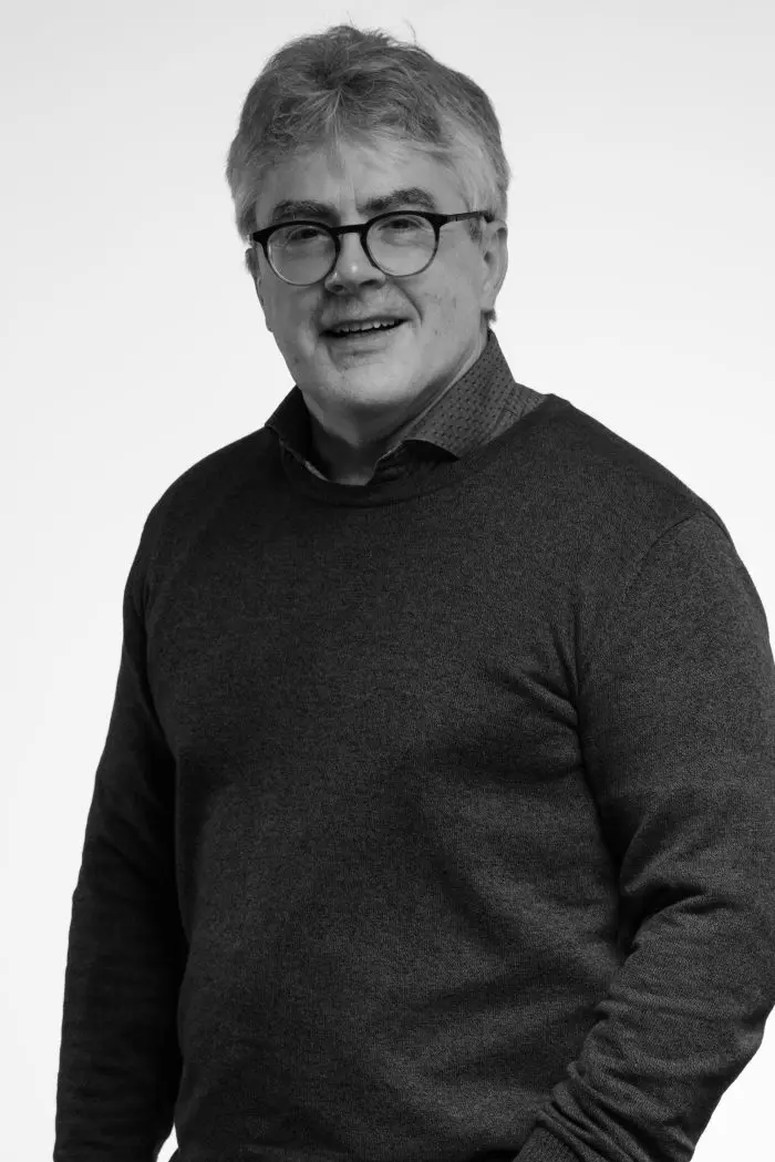Portrait en noir et blanc d'un homme d'âge moyen avec des lunettes, des cheveux gris et un léger sourire, portant un pull sombre sur une chemise à col, debout sur un fond clair uni.
