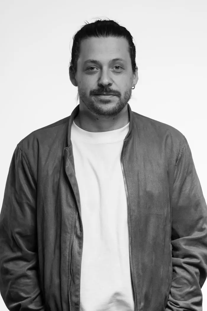 Un homme aux cheveux longs attachés, à la moustache et à la barbe, portant une veste légère sur une chemise blanche, se tient face à l'appareil photo sur un fond uni et clair. L'image est en noir et blanc.