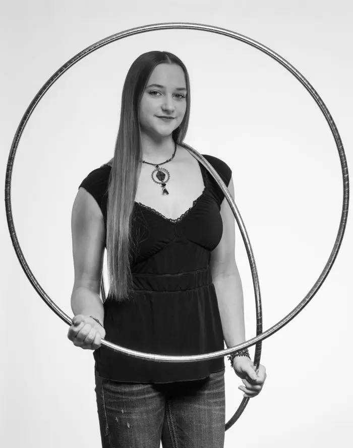 Une femme aux longs cheveux raides se tient debout, tenant un grand cerceau verticalement devant elle. Elle porte un haut noir, un jean et un collier. L'image est en noir et blanc sur un fond uni.