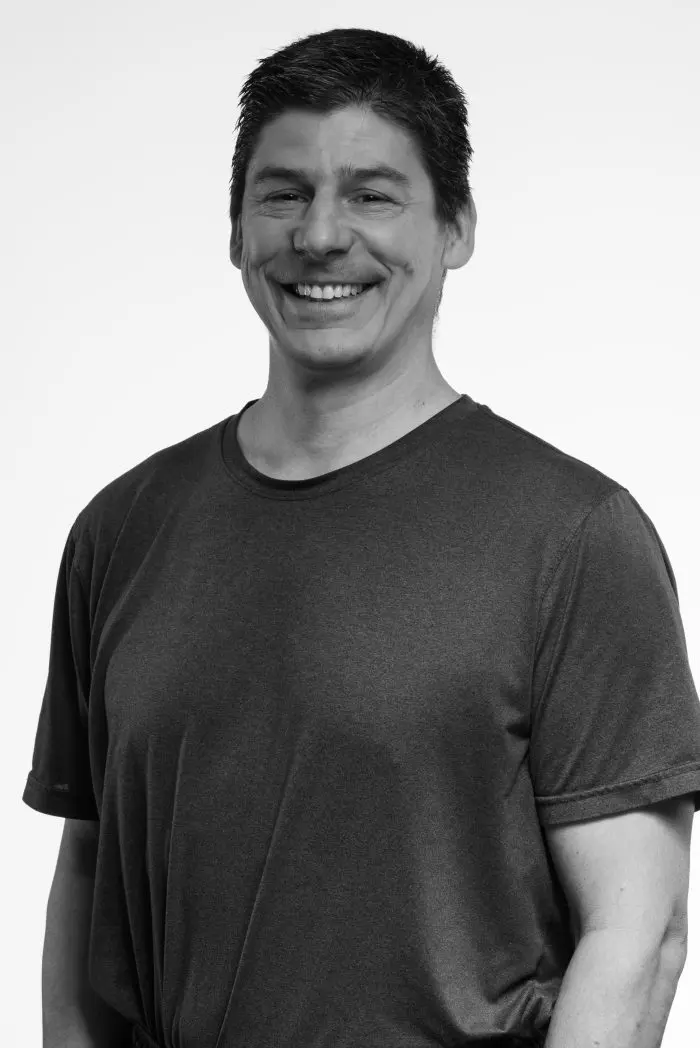 Un homme aux cheveux courts et foncés, vêtu d'un tee-shirt foncé, sourit sur un fond clair et uni. L'image est en noir et blanc.