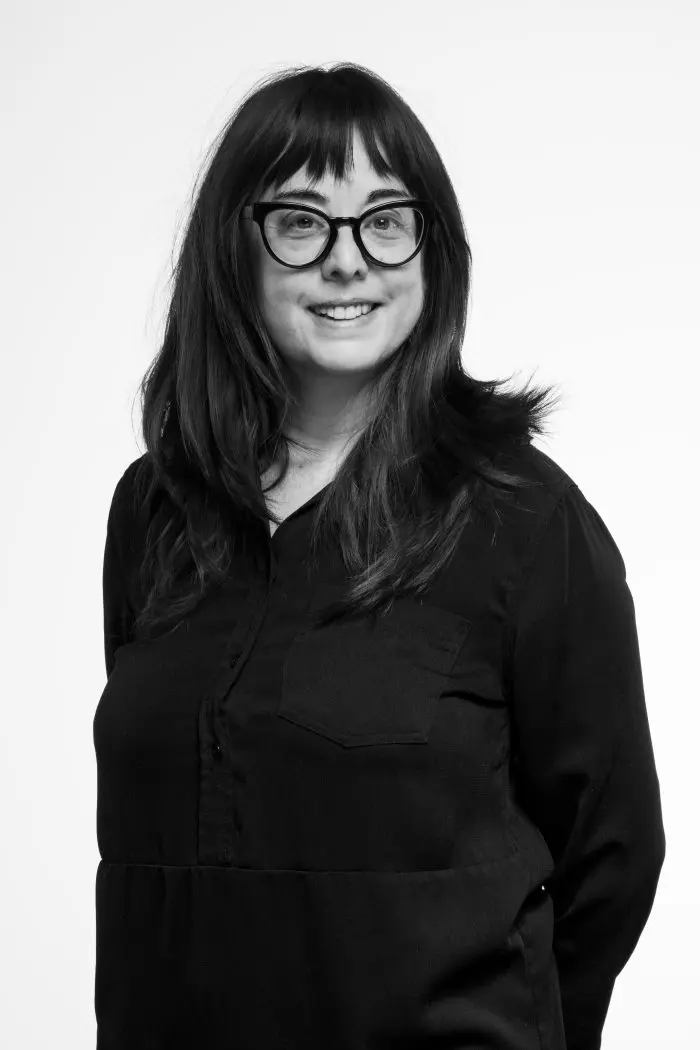 Une femme aux longs cheveux noirs et aux lunettes, vêtue d'une chemise sombre à manches longues, sourit sur un fond blanc uni.