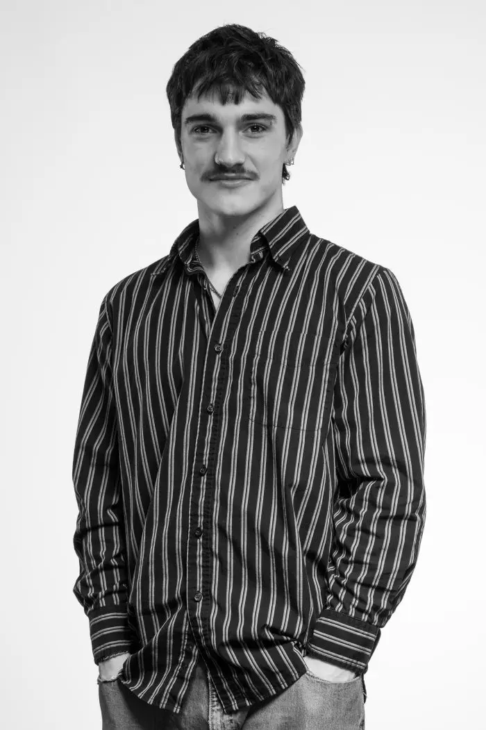 Un jeune homme moustachu se tient debout, les mains dans les poches, vêtu d'une chemise boutonnée rayée et d'un jean, sur un fond blanc uni.
