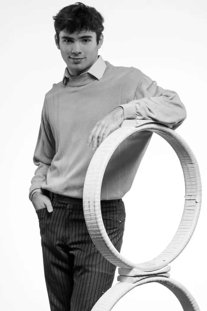 Un jeune homme vêtu d'un pull léger et d'un pantalon rayé se tient debout, une main dans la poche, appuyé sur deux cerceaux circulaires en osier empilés, sur un fond blanc uni.