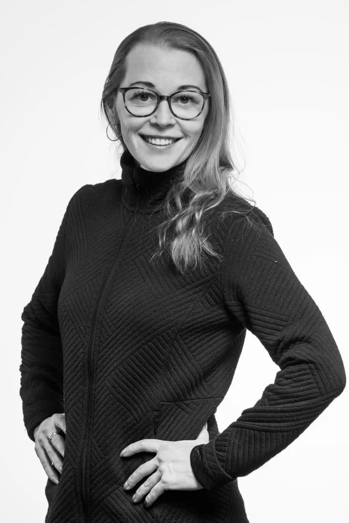 Une femme aux cheveux longs et clairs et aux lunettes sourit à l'appareil photo. Elle porte une veste sombre texturée et a les mains sur les hanches sur un fond clair et uni. L'image est en noir et blanc.