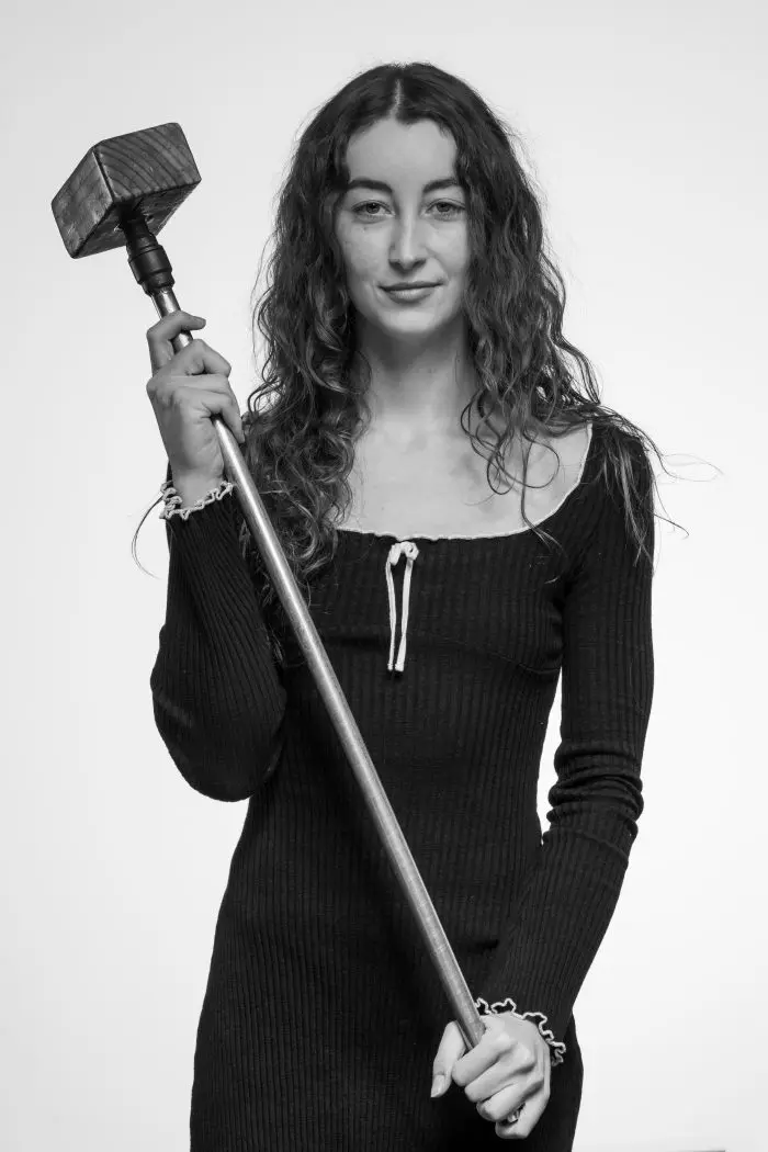 Une femme aux longs cheveux bouclés, vêtue d'une robe à manches longues, tient un gros marteau à la verticale sur son épaule, regardant l'appareil photo avec assurance sur un fond clair et uni.