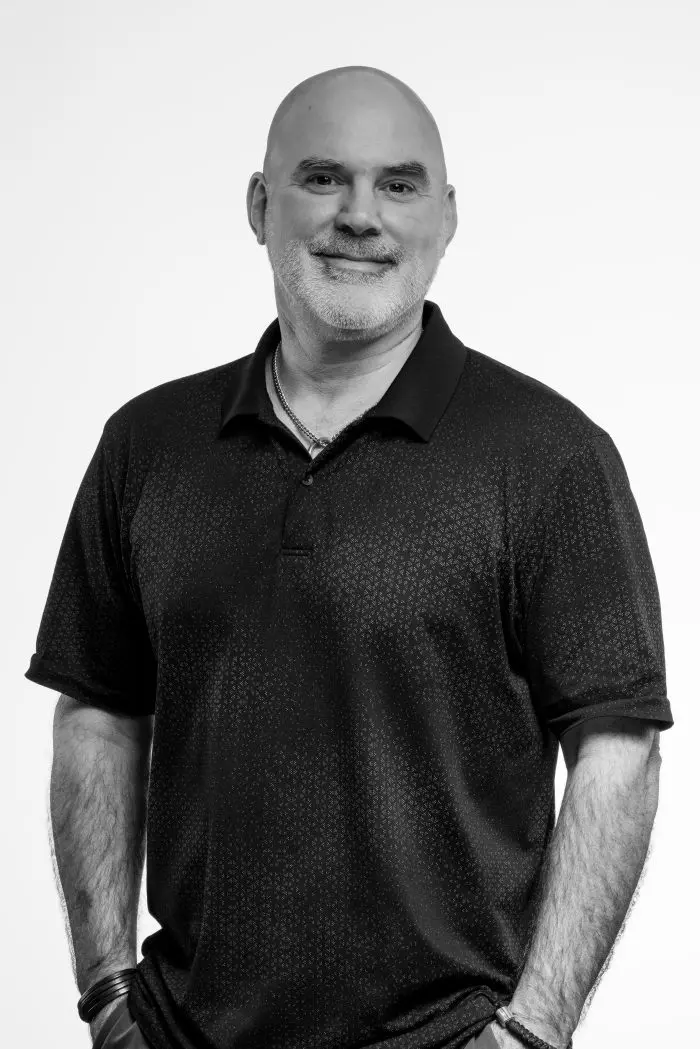 Un homme au crâne chauve et à la barbe courte, vêtu d'un polo sombre à manches courtes, se tient debout, les mains dans les poches, et sourit à l'appareil photo sur un fond blanc uni.