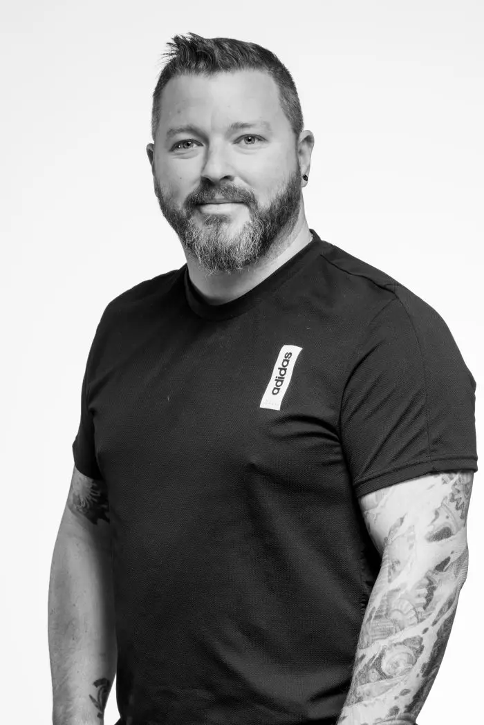Un homme aux cheveux courts et à la barbe, vêtu d'un t-shirt Adidas noir, se tient face à la caméra. Il a des tatouages sur les bras et sourit légèrement. La photo est en noir et blanc sur un fond uni.