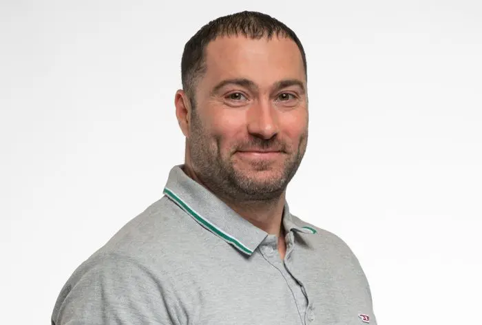 Un homme aux cheveux bruns courts et à la barbe sourit légèrement. Il porte un polo gris clair dont le col est orné de vert et de blanc. Il se tient debout sur un fond blanc uni.