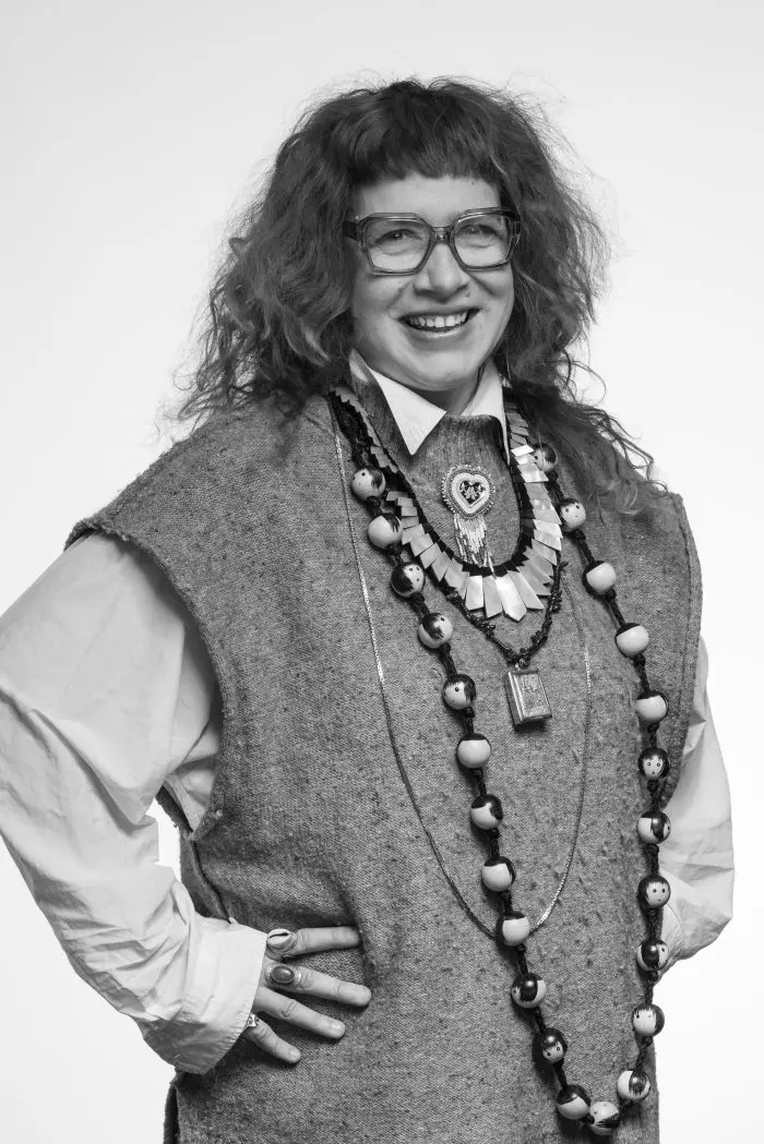 Une personne aux cheveux bouclés et aux lunettes sourit, portant un gilet texturé sur une chemise à col. Elle porte de nombreux colliers et bagues de grande taille et se tient debout, une main sur la hanche, sur un fond uni. La photo est en noir et blanc.