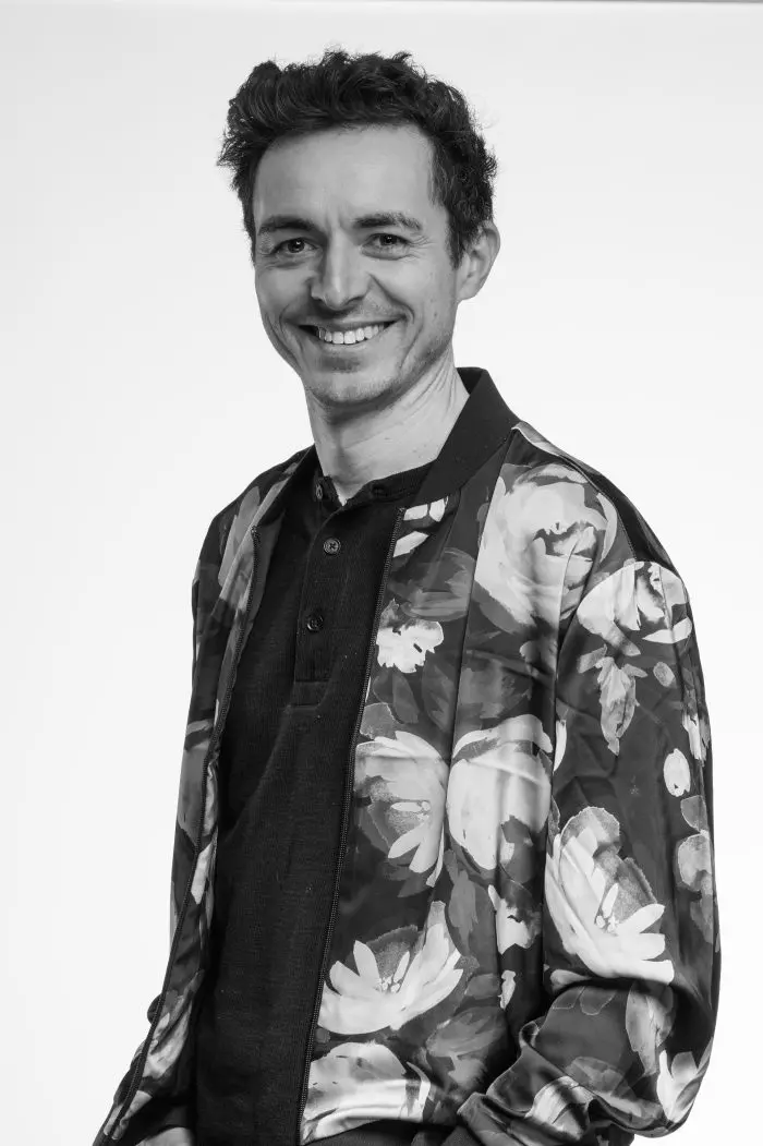Un homme aux cheveux courts et bouclés sourit à la caméra. Il porte une chemise sombre et une veste à motifs floraux. La photo est en noir et blanc sur un fond clair et uni.