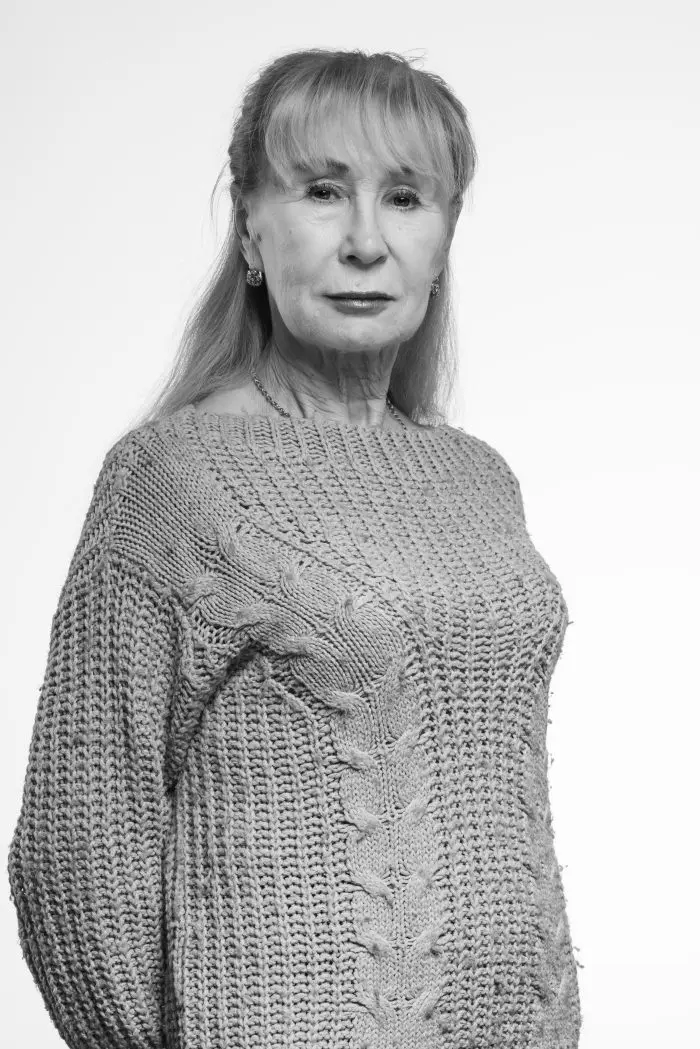 Portrait en noir et blanc d'une femme âgée aux cheveux longs, portant un pull en tricot texturé. Elle est légèrement tournée vers le côté et regarde directement l'appareil photo avec une expression neutre. L'arrière-plan est simple et clair.