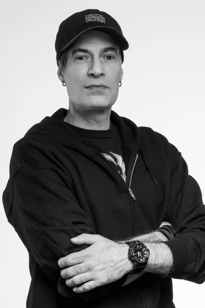 Une personne portant un sweat à capuche noir, une casquette noire et une montre-bracelet pose les bras croisés sur un fond uni. Elle a les cheveux courts, des boucles d'oreilles et une expression faciale neutre.