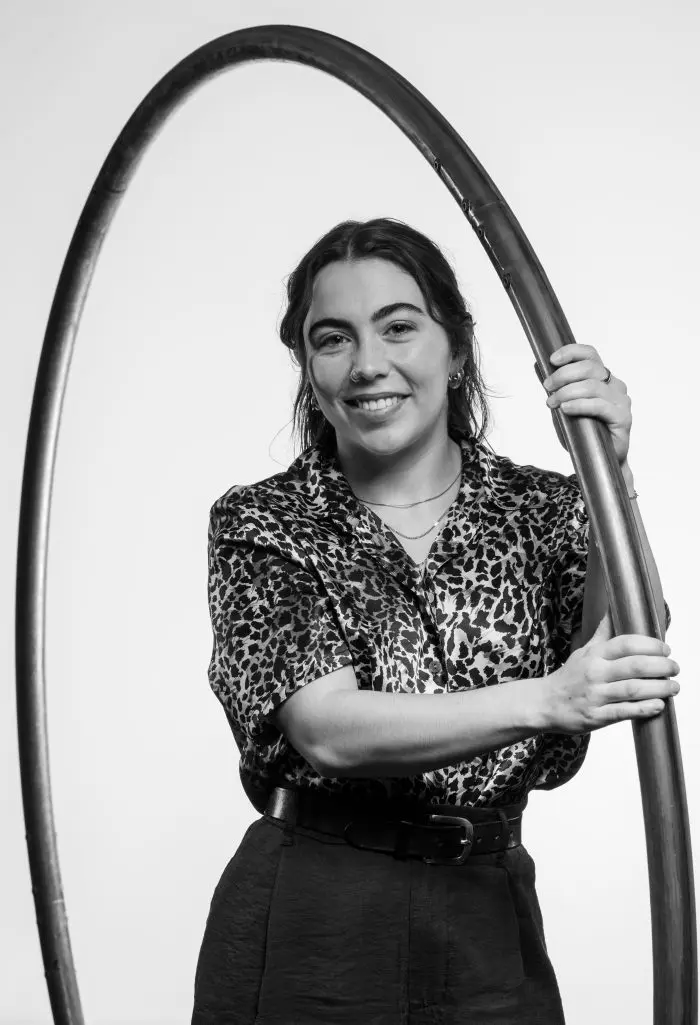 Une femme portant une chemise à imprimé léopard et un pantalon sombre sourit tout en tenant un grand cerceau métallique à la verticale sur un fond uni.