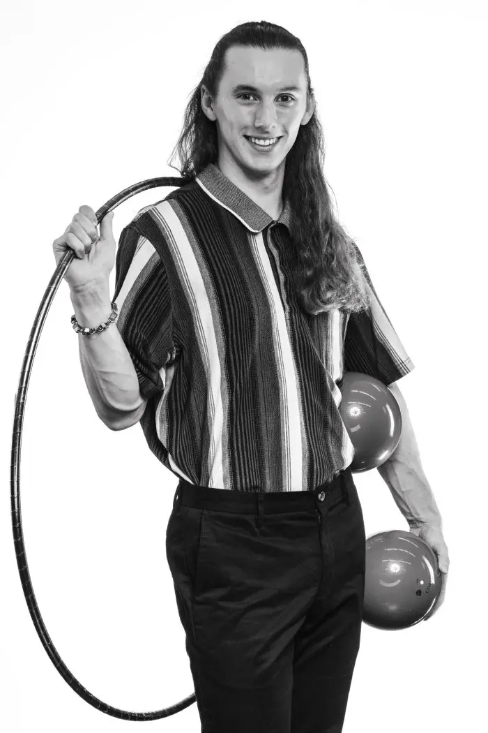 Une personne aux cheveux longs, vêtue d'une chemise rayée et d'un pantalon sombre, sourit en tenant un hula hoop dans une main et deux balles de jonglage dans l'autre. La photo est en noir et blanc.