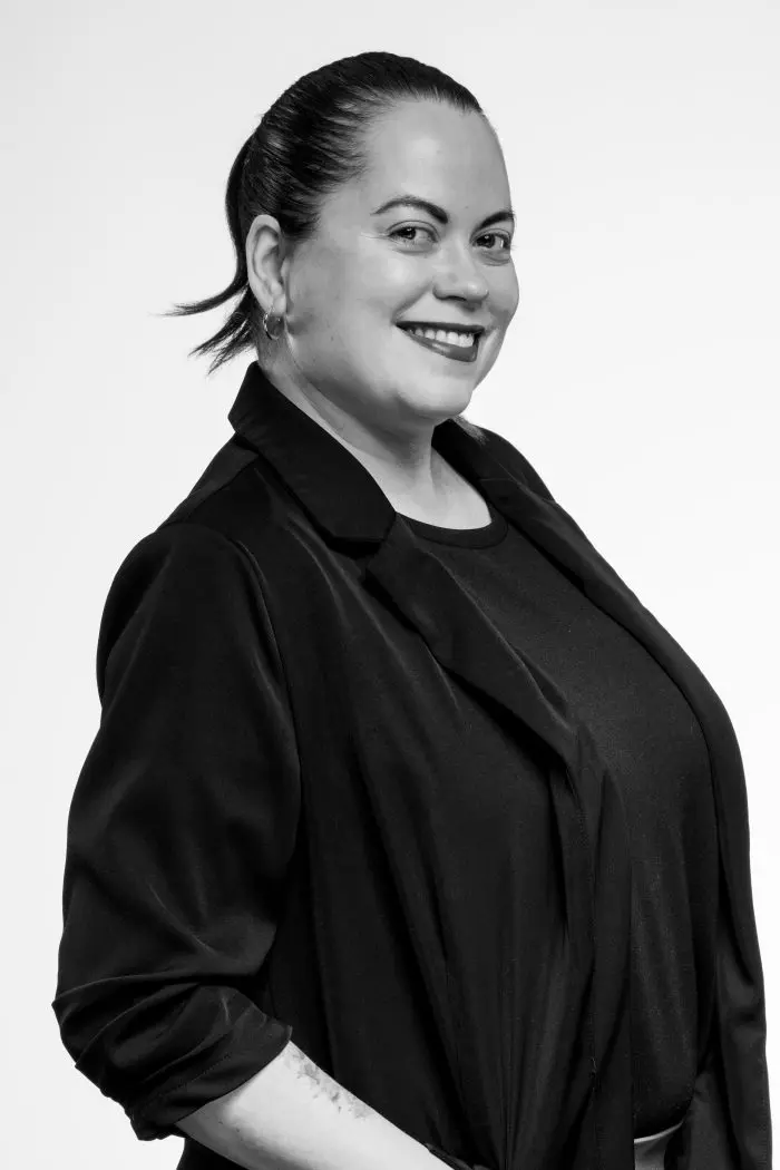 Une femme aux cheveux noirs attachés, portant un blazer noir sur un haut sombre, sourit sur un fond blanc uni. La photo est en noir et blanc.