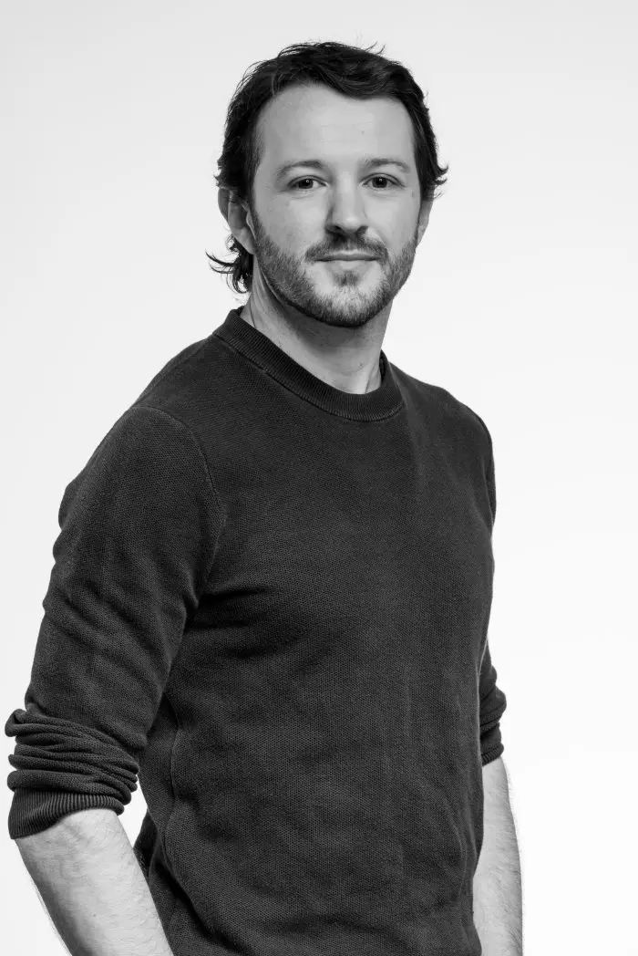 Un homme aux cheveux bruns mi-longs et à la barbe, vêtu d'un pull sombre, se tient face à la caméra avec un léger sourire sur un fond blanc uni.