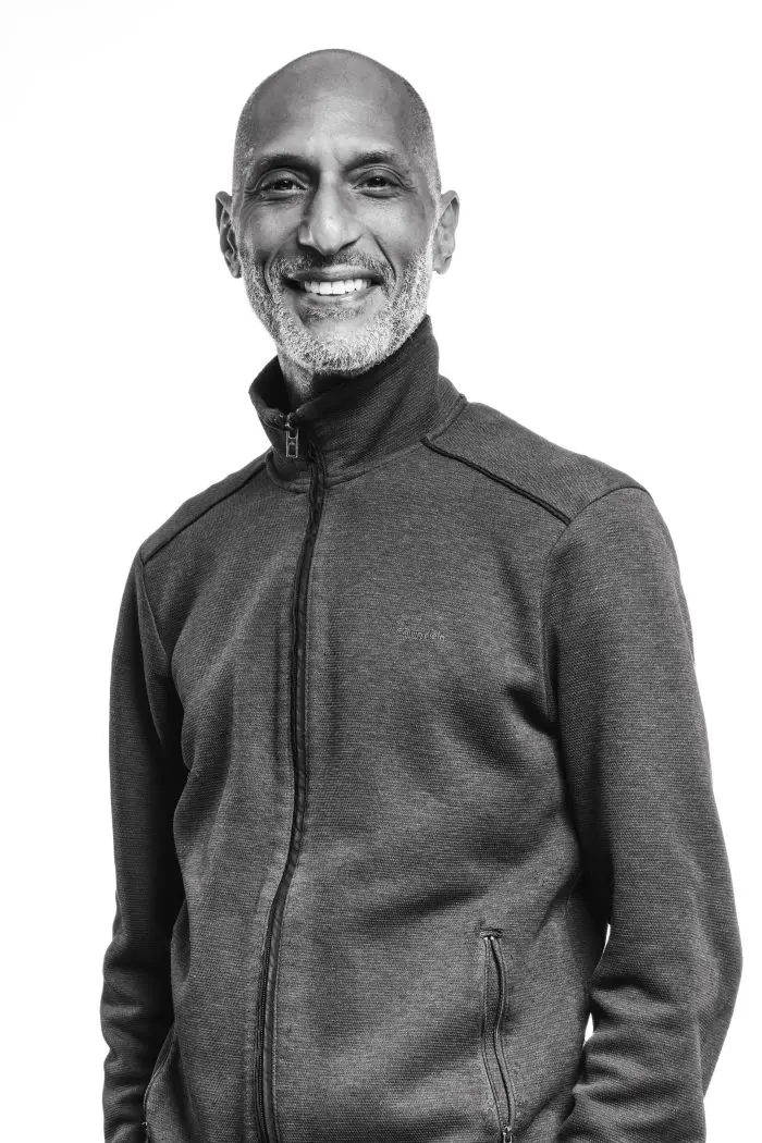 Un homme souriant, au crâne rasé et à la courte barbe grise, vêtu d'une veste sombre à fermeture éclair, se tient sur un fond blanc uni.