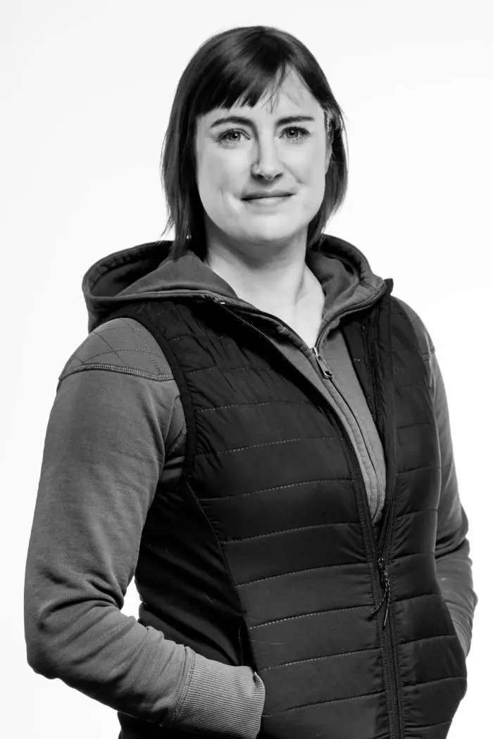 Une femme aux cheveux courts, vêtue d'un sweat à capuche et d'un gilet matelassé, se tient debout, une main dans la poche, regardant l'appareil photo. L'image est en noir et blanc sur un fond uni.