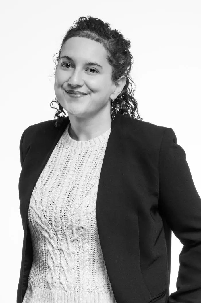 Portrait en noir et blanc d'une personne aux cheveux bouclés, portant un pull en maille clair et un blazer foncé, souriant et regardant légèrement de côté sur un fond uni.