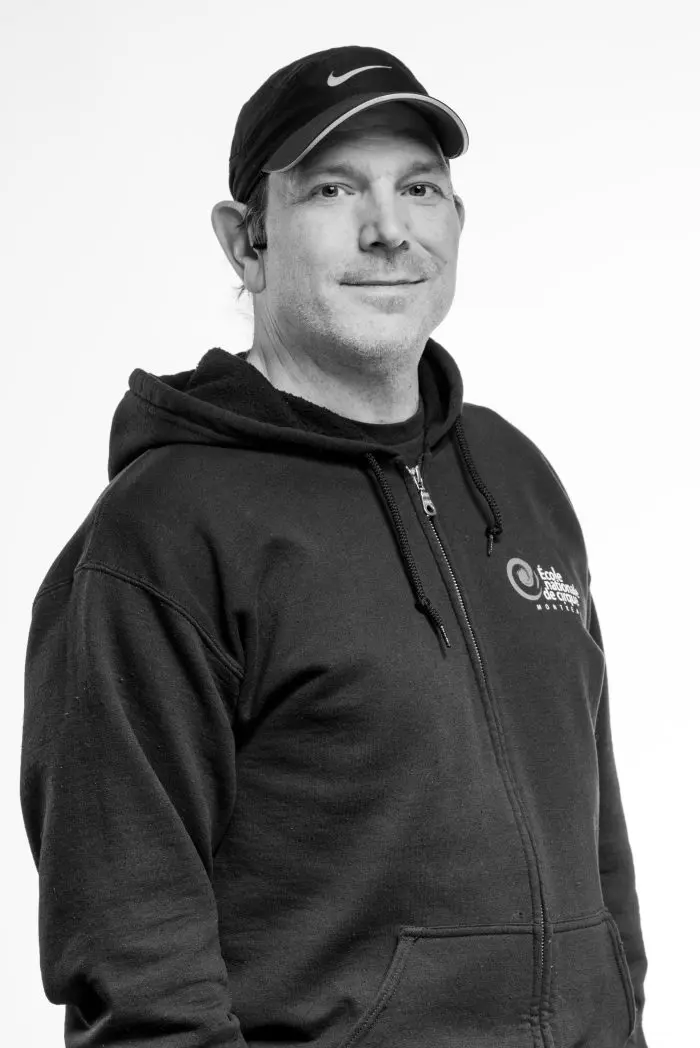 Un homme portant un sweat à capuche sombre et une casquette Nike noire, souriant légèrement tout en regardant la caméra. L'image est en noir et blanc sur fond clair.