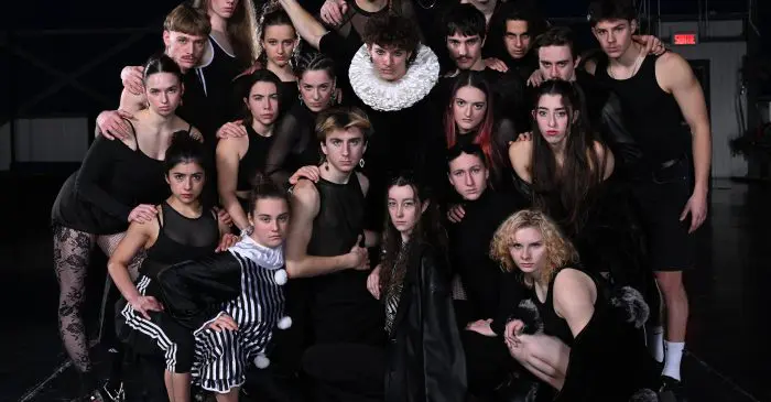 Un groupe de jeunes adultes aux costumes sombres et dramatiques et aux expressions intenses posent ensemble sur scène pour leur spectacle des finissants ; l'un d'eux tient une épingle de jonglage tandis qu'un autre arbore un col à volants, tous regardant directement l'appareil photo.