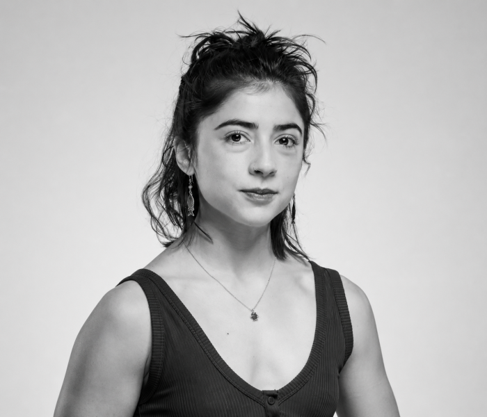 Portrait en noir et blanc de Metztli Albalat, une jeune femme aux cheveux foncés attachés, vêtue d'un haut sans manches, d'un collier et de boucles d'oreilles, qui regarde calmement l'objectif sur un fond uni.