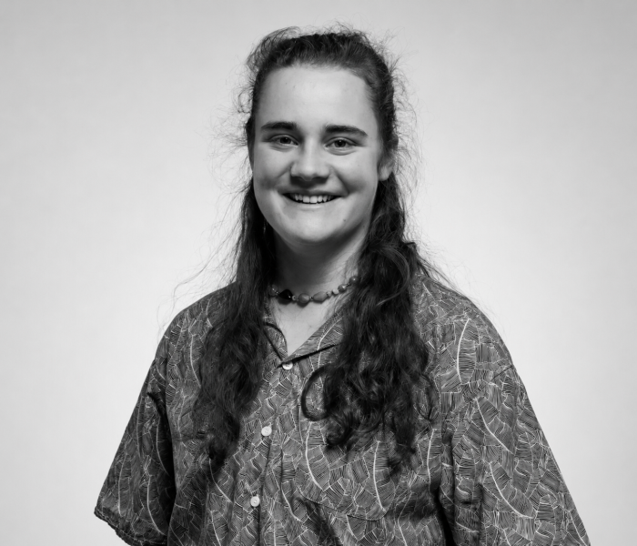Zora Eichenberger, aux longs cheveux bruns, sourit à l'appareil photo. Elle porte une chemise à motifs et un collier de perles. La photo en noir et blanc est prise sur un fond uni.