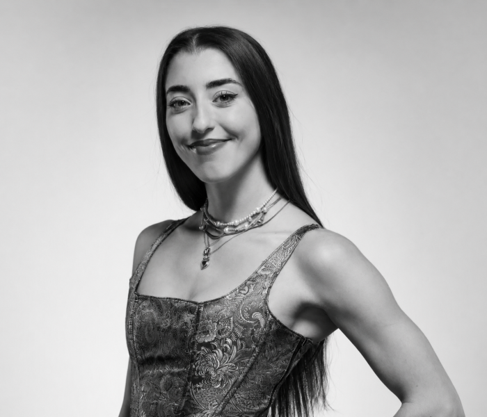 Martina Salza, une jeune femme aux longs cheveux bruns, sourit dans une robe sans manches à motifs et plusieurs colliers superposés, posant avec assurance devant un fond uni dans ce portrait en noir et blanc.