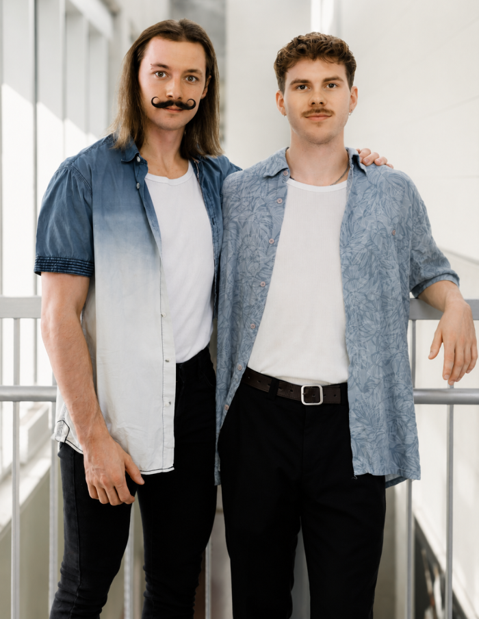 Colin Vuilleme et Nino Bartolini, tous deux moustachus, se tiennent côte à côte à l'intérieur, vêtus de chemises blanches assorties et de chemises bleues boutonnées et ouvertes. L'un d'eux pose son bras sur l'épaule de l'autre dans un espace lumineux et moderne doté de grandes baies vitrées.