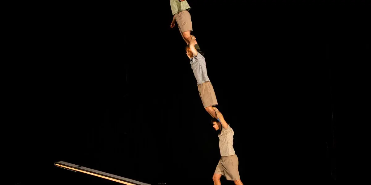 Trois acrobates de TriSwiss exécutent un numéro d'équilibre dans un décor sombre ; l'un d'eux se tient sur une balançoire à bascule et tient un autre acrobate, qui soutient une troisième personne debout sur ses épaules.