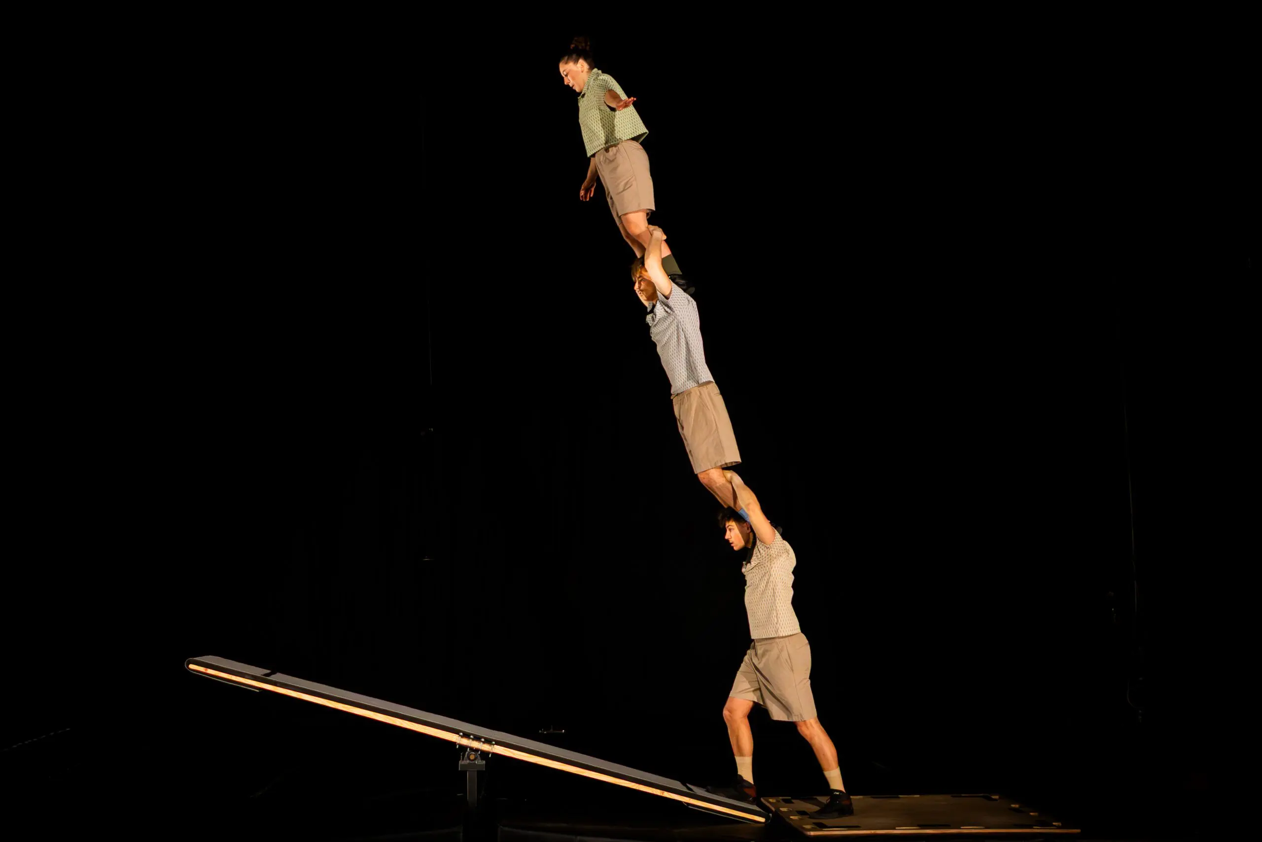 Trois acrobates de TriSwiss exécutent un numéro d'équilibre dans un décor sombre ; l'un d'eux se tient sur une balançoire à bascule et tient un autre acrobate, qui soutient une troisième personne debout sur ses épaules.