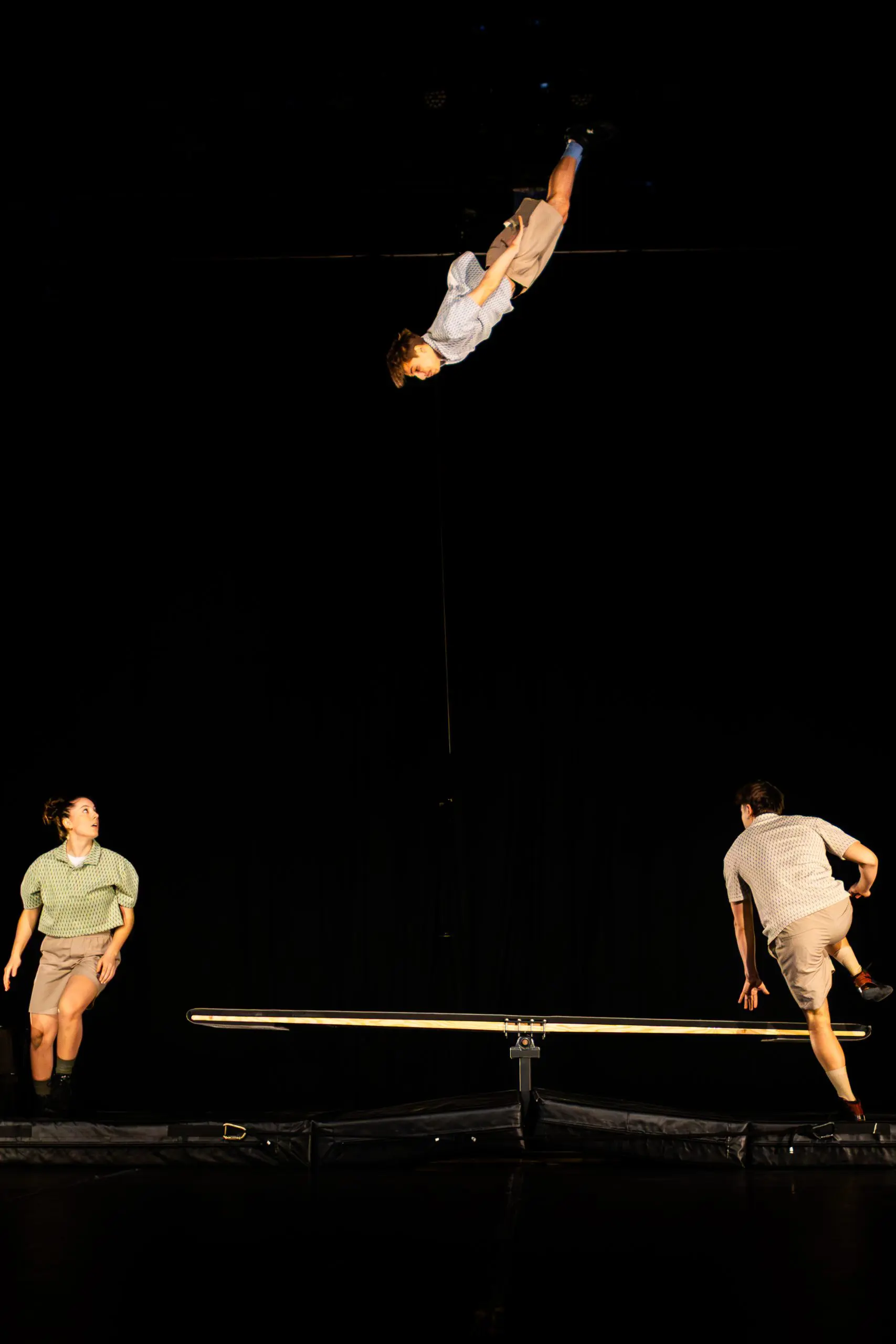 Trois artistes TriSwiss en tenue décontractée sont sur scène avec une planche à bascule. L'un d'eux est en l'air au milieu d'un saut tandis que les deux autres le regardent de chaque côté, le tout sur un fond sombre qui met en valeur l'action palpitante.