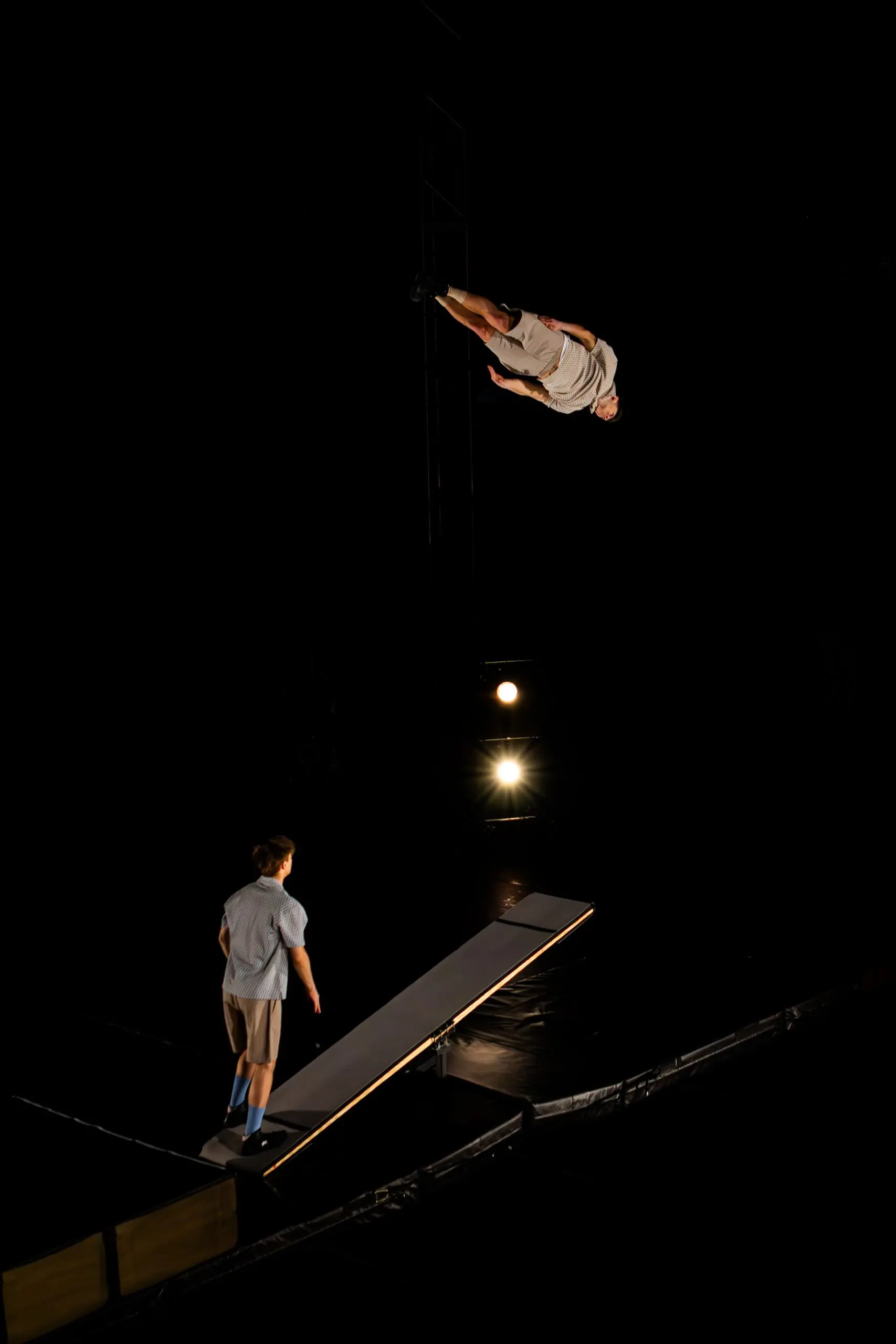 Un athlète de TriSwiss exécute un saut périlleux arrière en l'air au-dessus d'un appareil ressemblant à une balançoire, tandis qu'une autre personne se tient en dessous, regardant vers le haut, le tout sur un fond sombre avec un éclairage dramatique.
