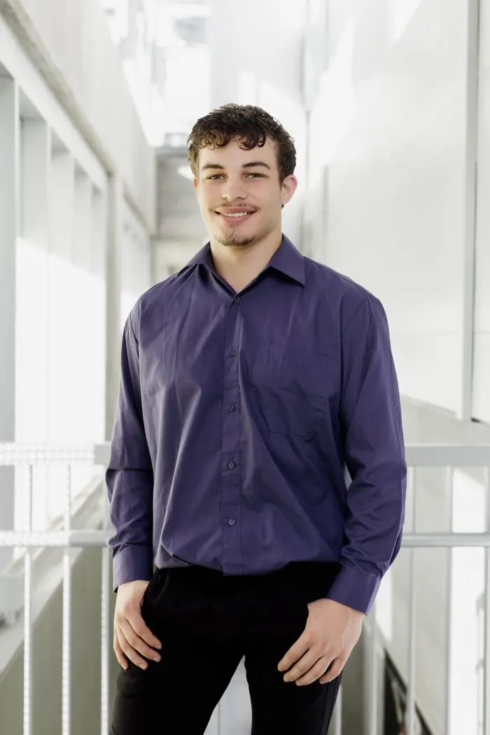 Kayden Woodridge, un jeune homme aux cheveux courts et bouclés et à la barbe, se tient à l'intérieur en souriant. Il porte une chemise à boutons violette et un pantalon noir, la main gauche dans la poche. L'arrière-plan lumineux présente des murs et des fenêtres blancs.