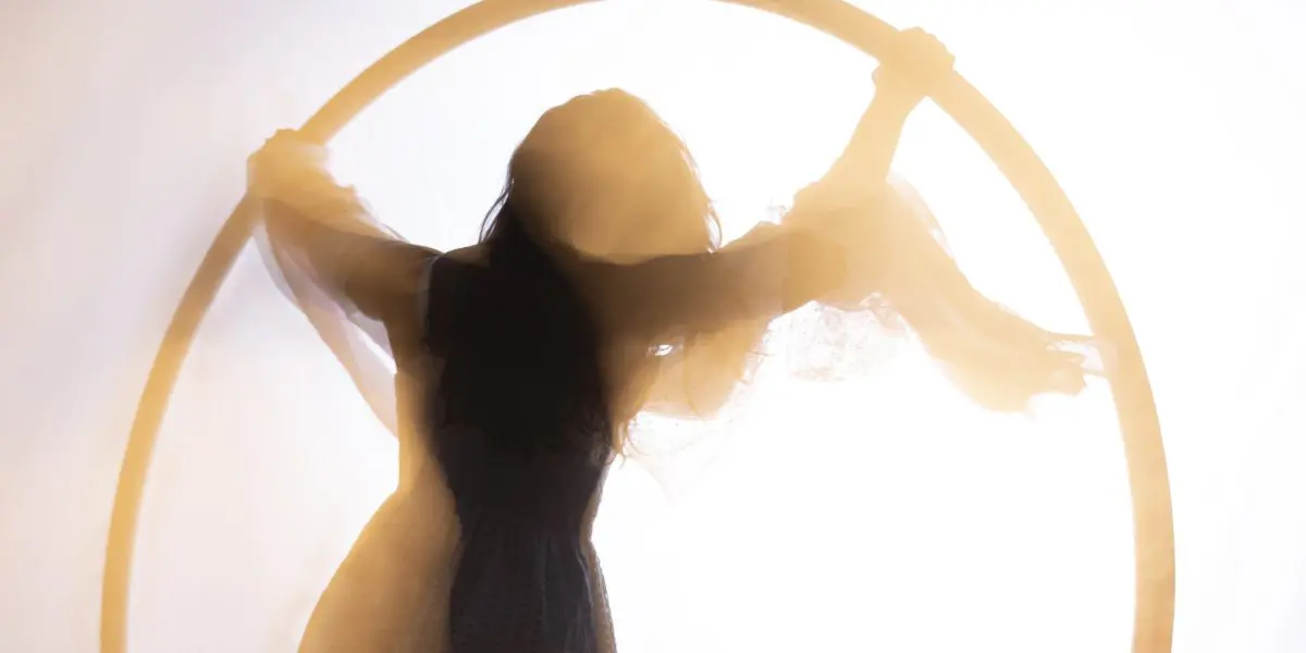 Silhouette d'une personne en robe tenant un grand cerceau circulaire, avec un tissu transparent drapé sur les bras, sur un fond lumineux, évoquant l'art gracieux de Léonie Salcines.
