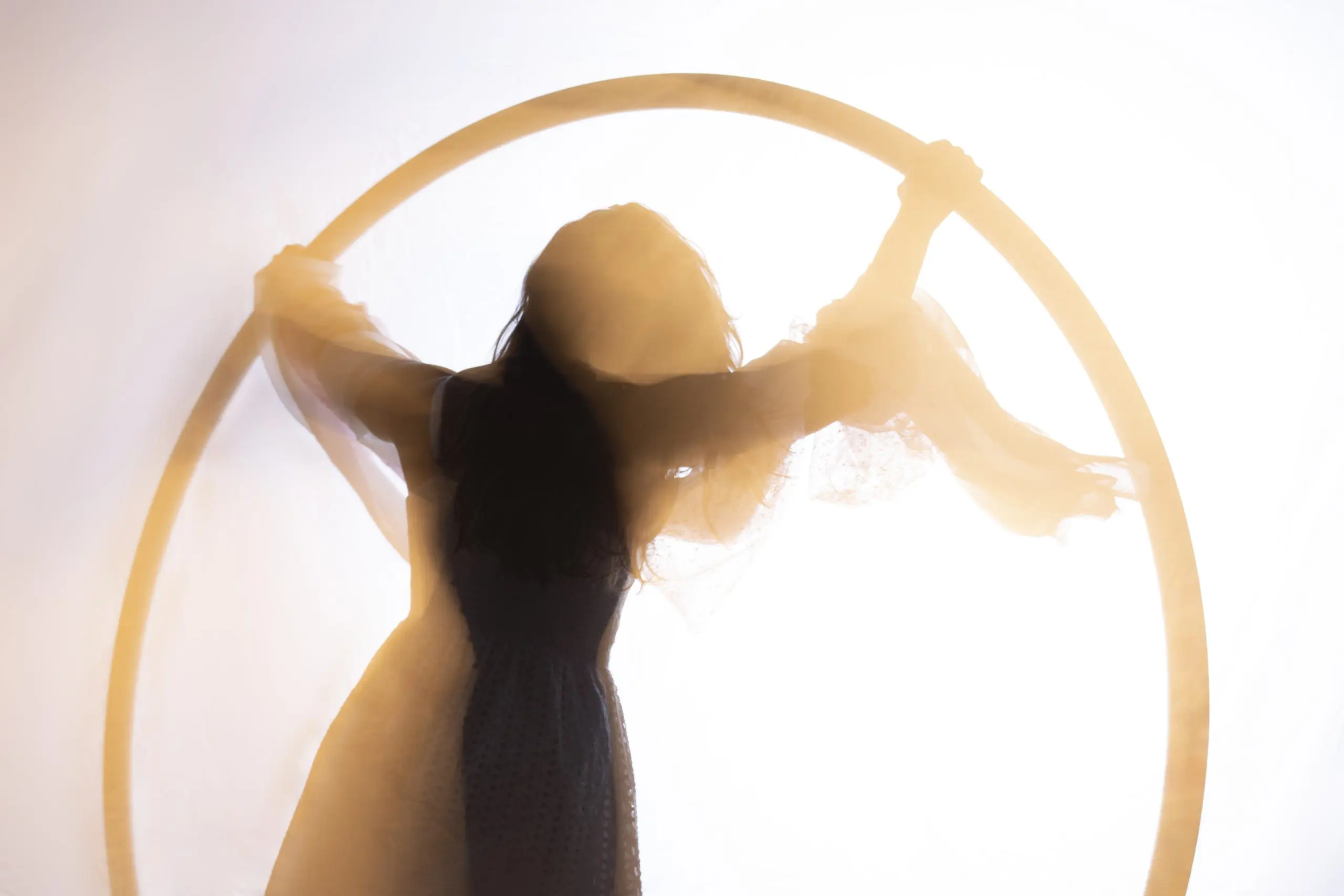 Silhouette d'une personne en robe tenant un grand cerceau circulaire, avec un tissu transparent drapé sur les bras, sur un fond lumineux, évoquant l'art gracieux de Léonie Salcines.