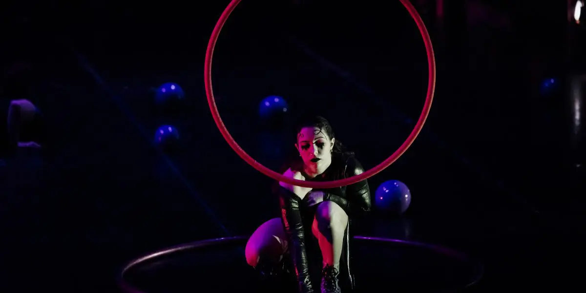 Léonie Salcines, vêtue de vêtements sombres, est accroupie de façon dramatique à l'intérieur d'un cerceau circulaire, tandis qu'un autre est posé sur le sol. Des balles bleues sont éparpillées sur la scène, et l'éclairage dramatique accentue son visage et sa forme.