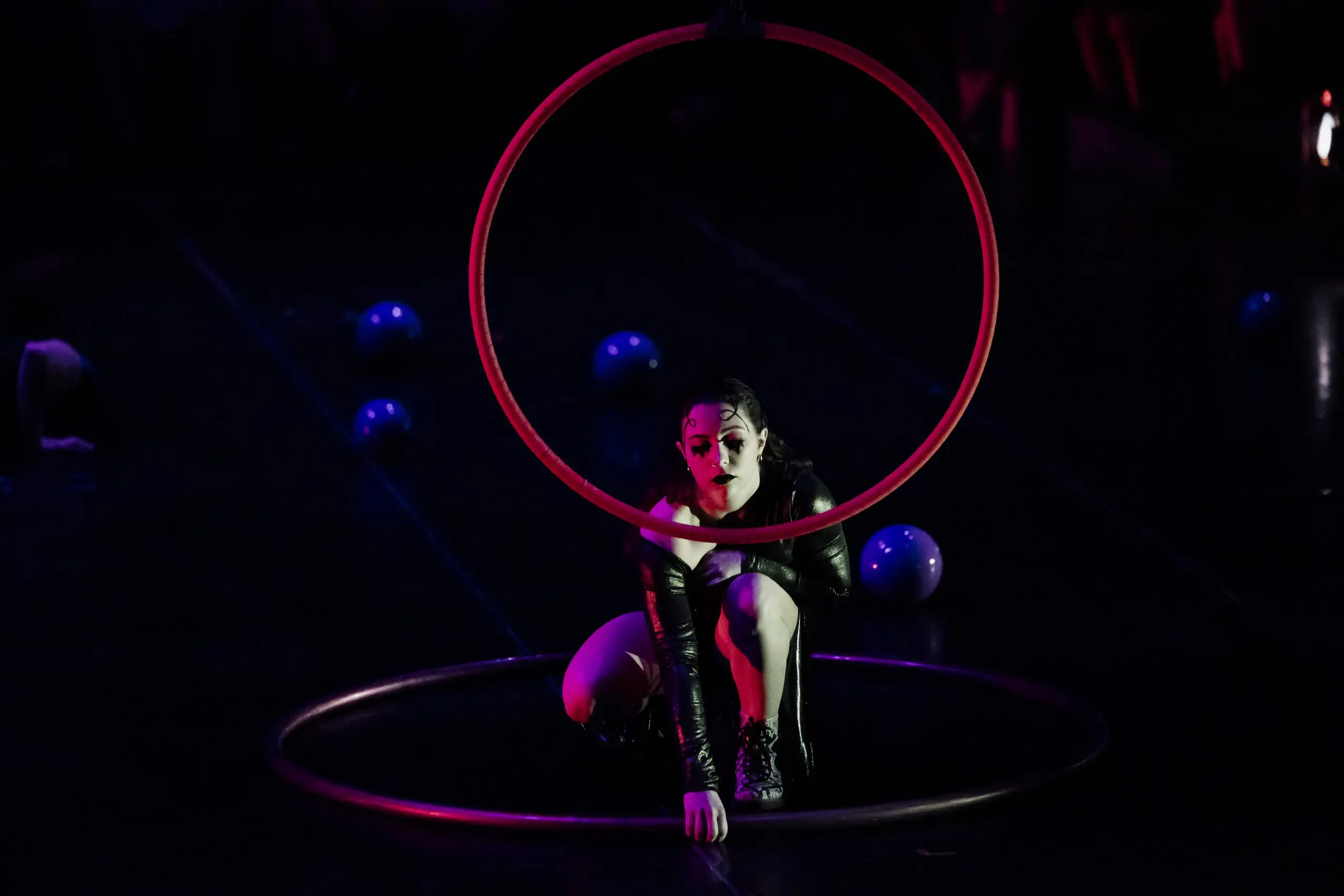 Léonie Salcines, vêtue de vêtements sombres, est accroupie de façon dramatique à l'intérieur d'un cerceau circulaire, tandis qu'un autre est posé sur le sol. Des balles bleues sont éparpillées sur la scène, et l'éclairage dramatique accentue son visage et sa forme.
