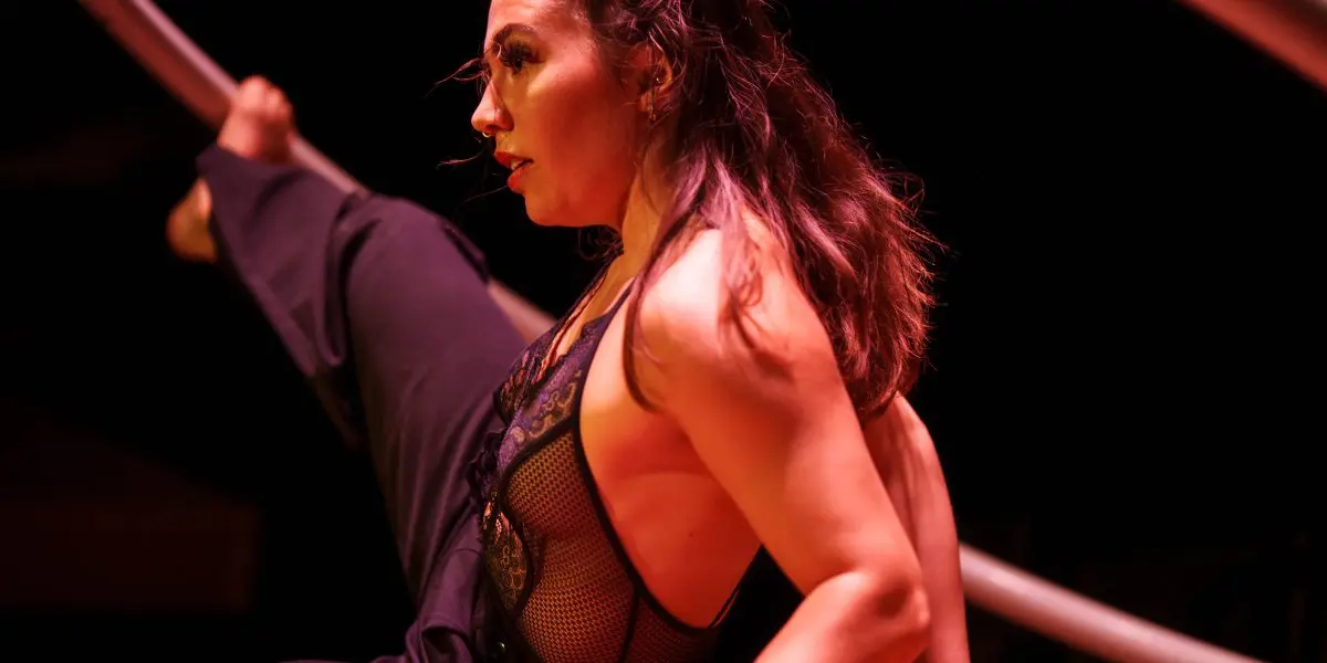 Léonie Salcines, vêtue d'une tenue en maille noire, prend une pose de contorsion flexible sur scène, tenant un grand cerceau métallique avec une jambe levée en hauteur sous un éclairage dramatique.